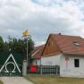 A house with red roof tiles, a green fence with the Alpha Omega symbol, two yellow flags and murals.