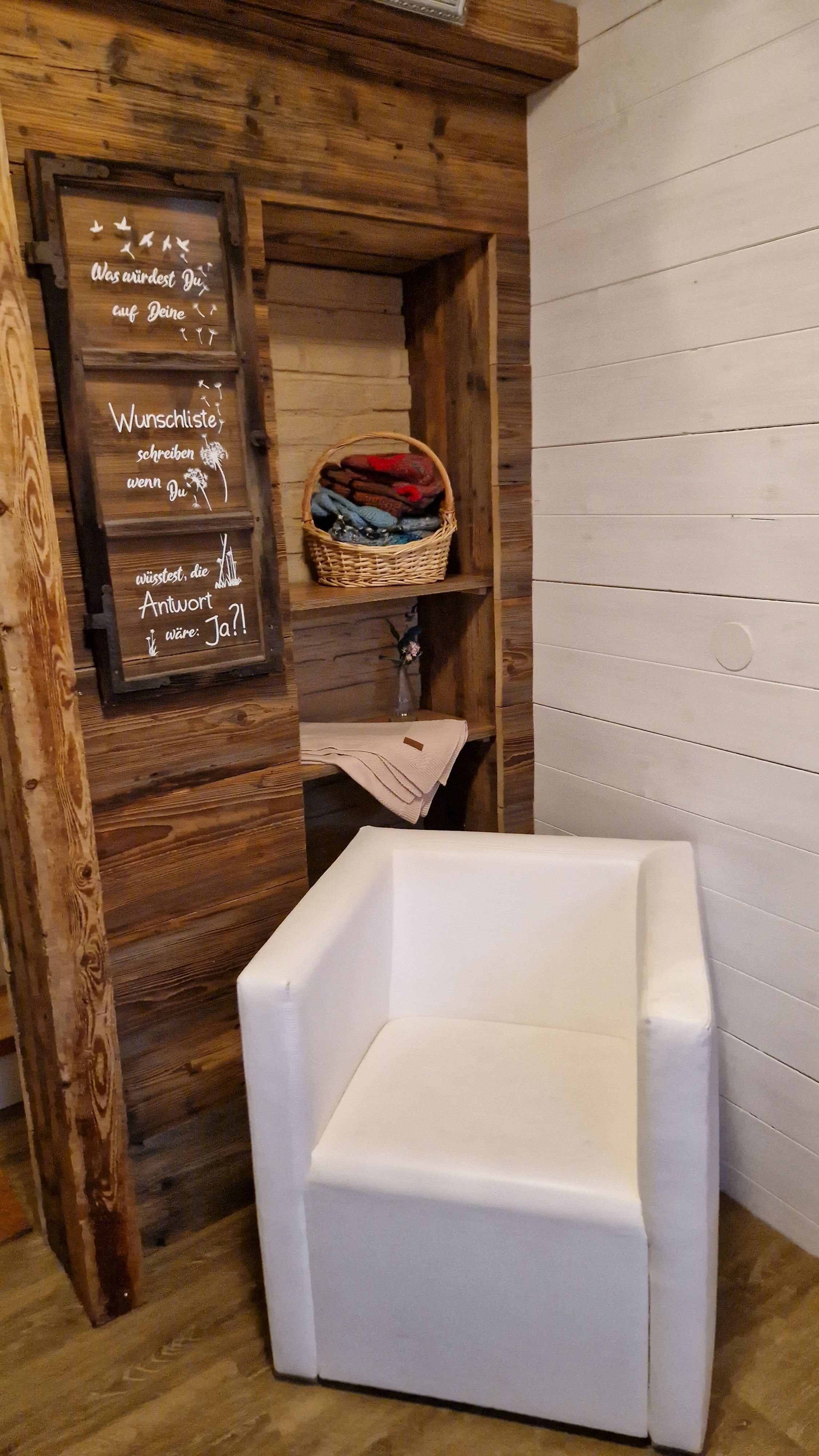 A cozy room with a white armchair, a shelf with folded blankets, and a chalkboard with an inspiring message.