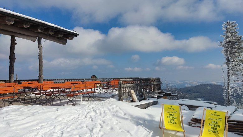 Snow-covered terrace with orange benches and two yellow deckchairs, surrounded by snow-covered trees and mountains in the background.