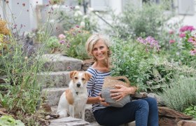 A woman sits with a dog on stone steps in a flowering garden.