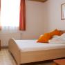 A simply furnished double room with a wooden bed, orange cushions and curtains, a chair and a picture on the wall.