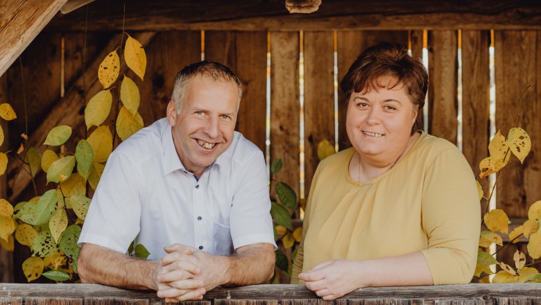 A man and a woman stand smiling by a wooden fence with autumn leaves in the background.