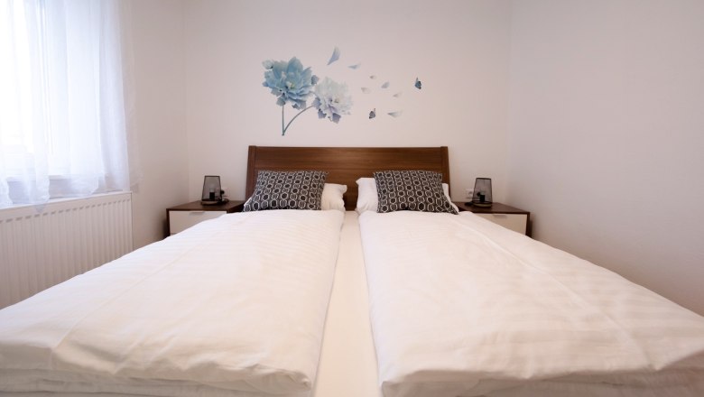 A modern bedroom with a double bed, white comforters and decorative pillows. There is a floral motif on the wall.