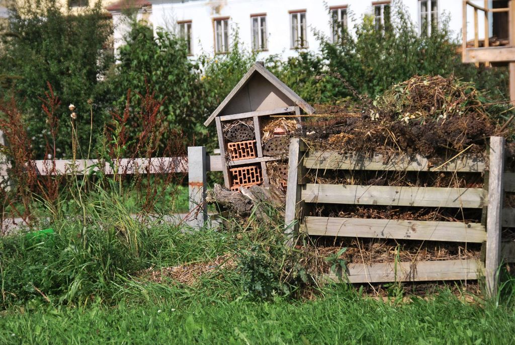 An insect hotel next to a compost heap in a garden with green grass and trees in the background.