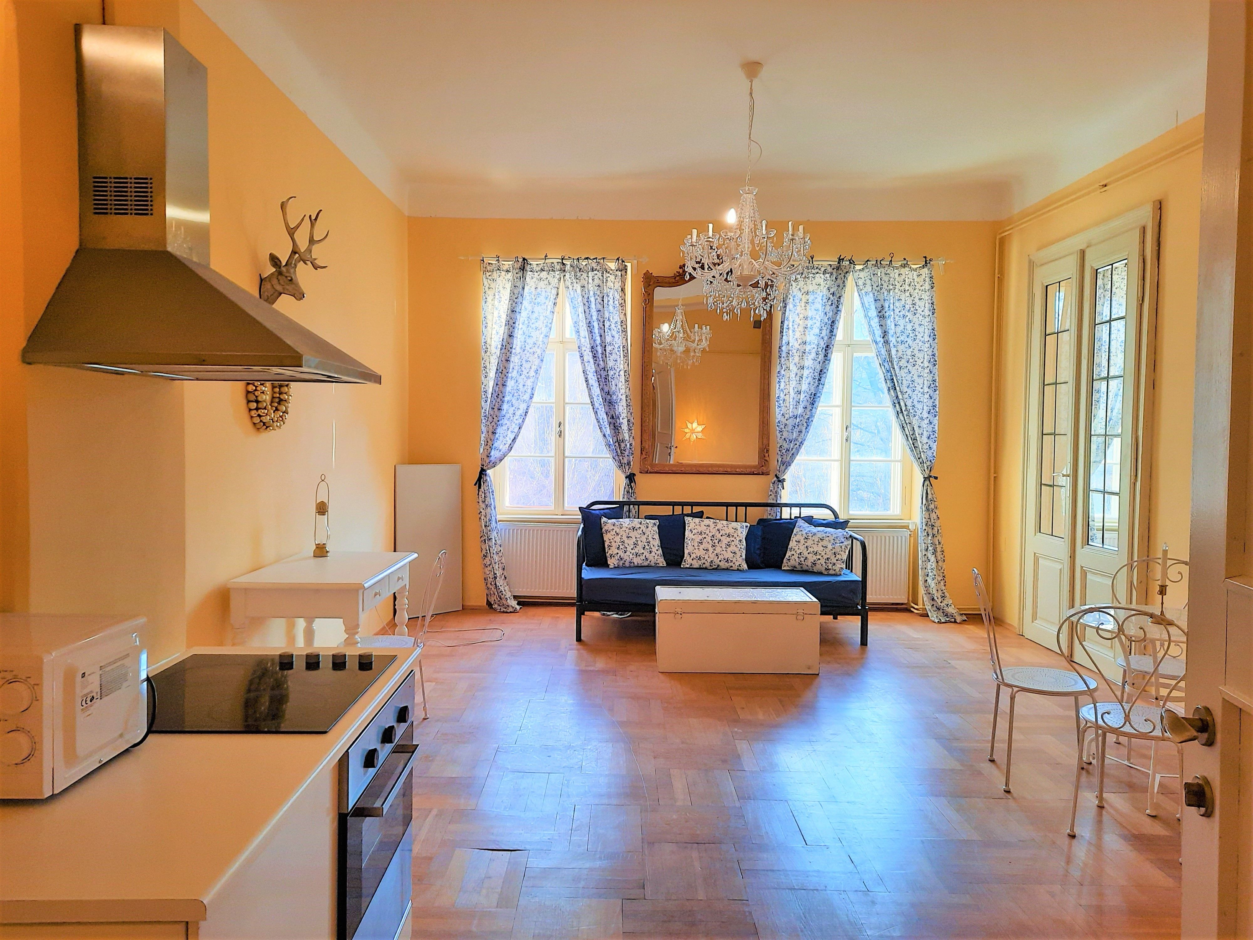 Living room with kitchen, dining table and sofa, yellow walls, chandelier, windows with curtains.