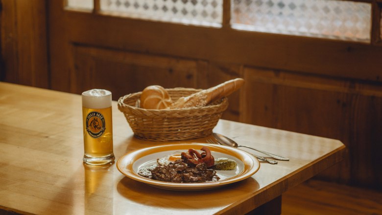 A plate with Fiaker goulash, fried egg and gherkin, next to it a glass of beer and a basket of bread on a wooden table.
