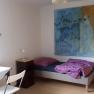 A simply furnished room with a bed, table, chair and large painting on the wall.