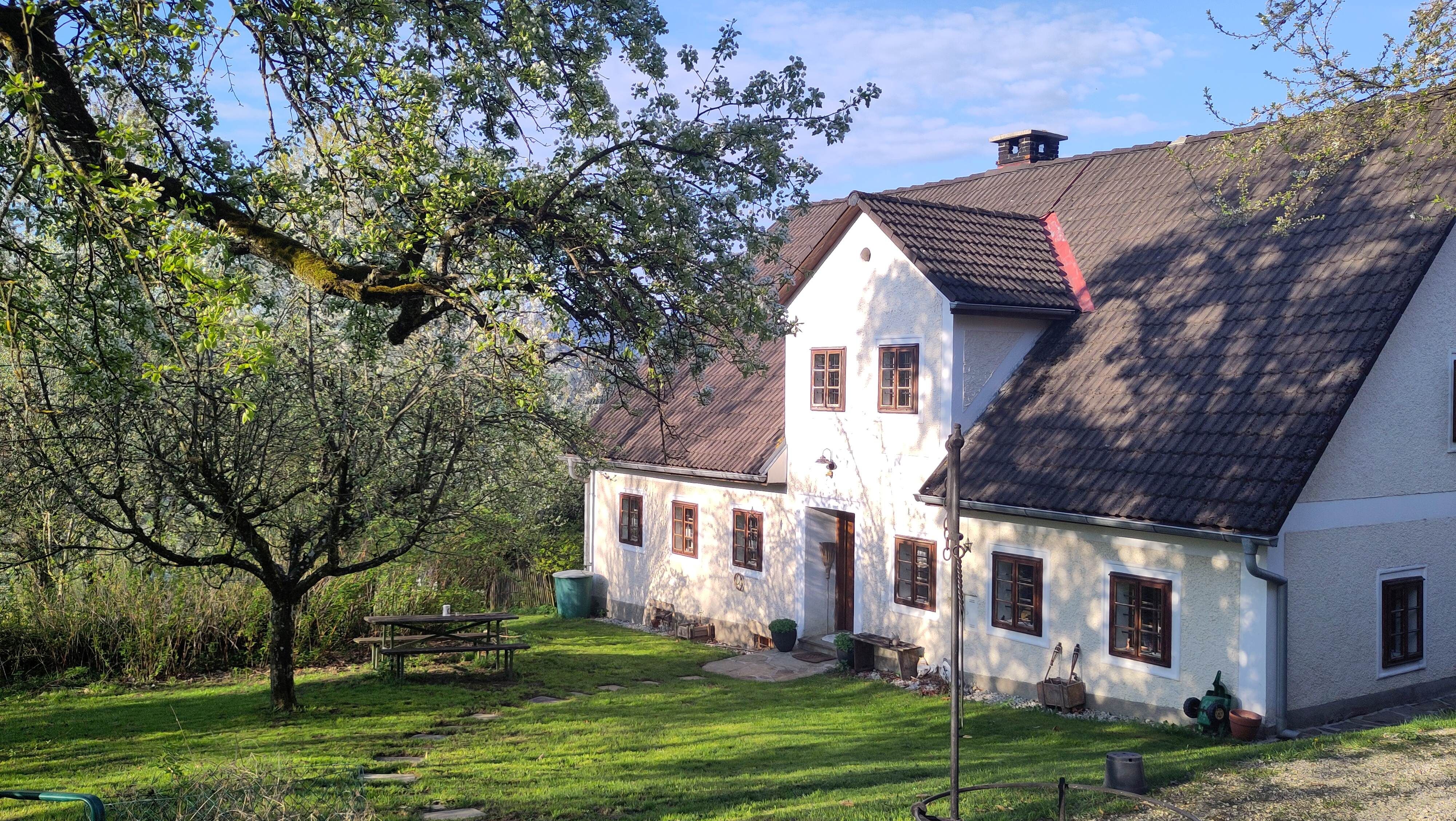 A traditional country house with white walls and brown windows, surrounded by green trees and a well-tended garden with a picnic table.