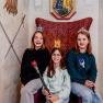 Three children with crowns and sceptres sit on a throne in a fairytale room.