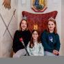 Three children with crowns and sceptres sit on a throne in a fairytale room.