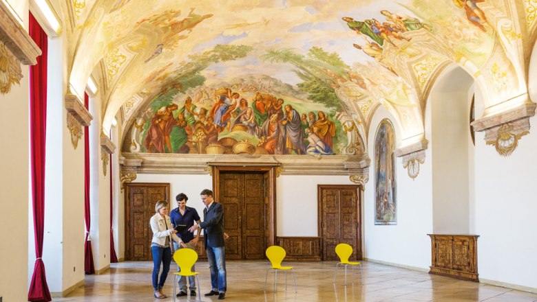 Interior view of a magnificent room in Göttweig Abbey with frescoes on the ceiling and three people talking.