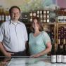 Two people stand in front of wine bottles and shelves of products in a winery.