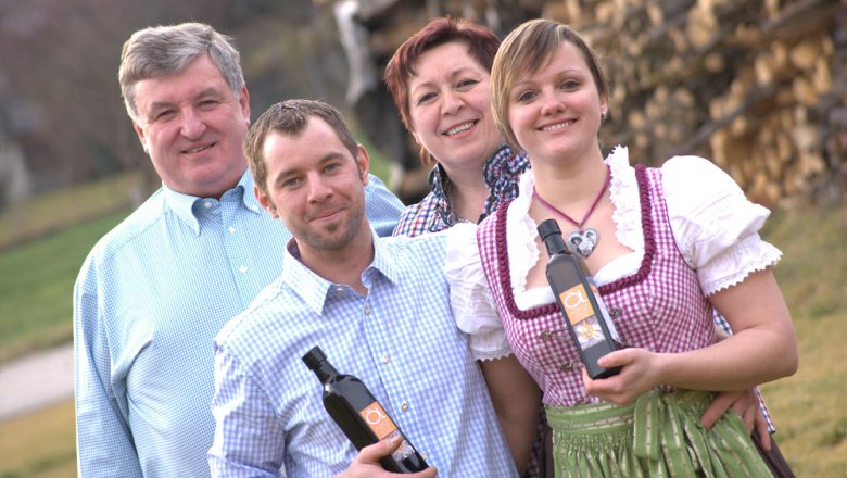 Four people are smiling at the camera, two are holding bottles of oil. A pile of wood can be seen in the background.