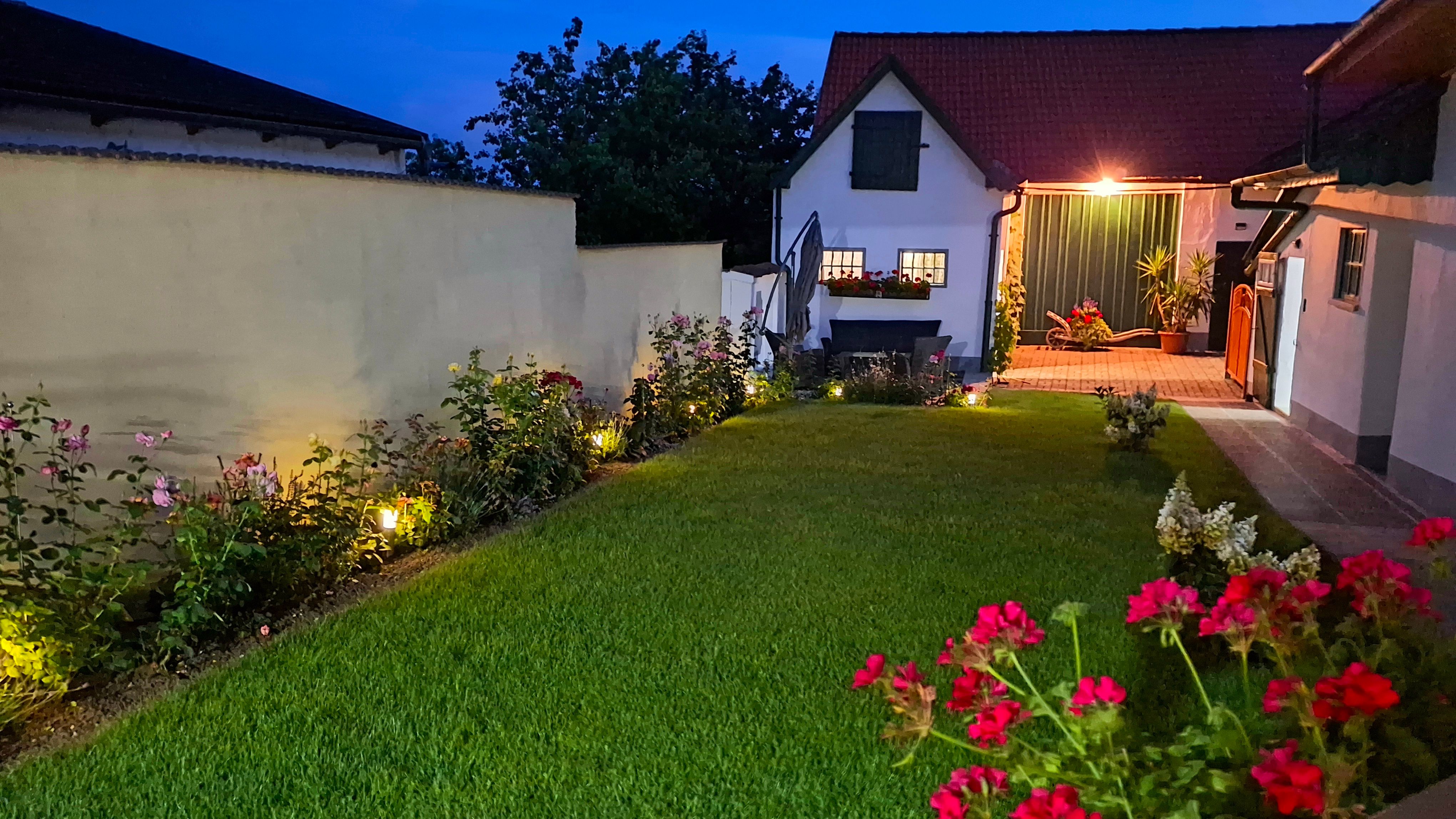 An illuminated courtyard at night with a manicured lawn, flower beds and a small building in the background.