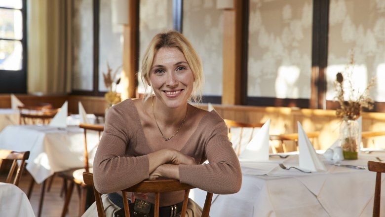 Woman sitting smiling in a restaurant with set tables.
