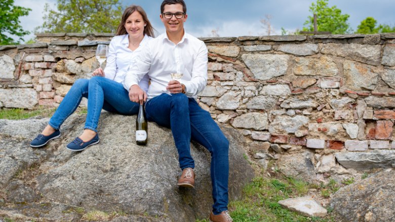 A man and a woman are sitting on a rock in front of a stone wall, holding wine glasses and a bottle of wine.
