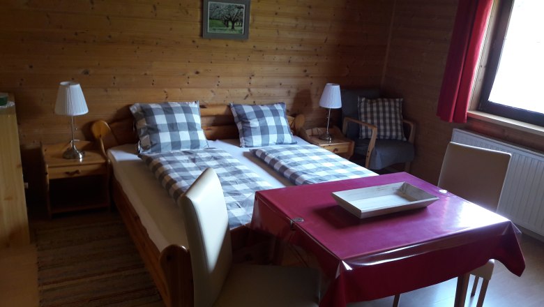 Cozy room with wooden walls, double bed, table with red tablecloth and chairs, lamp and window with red curtain.