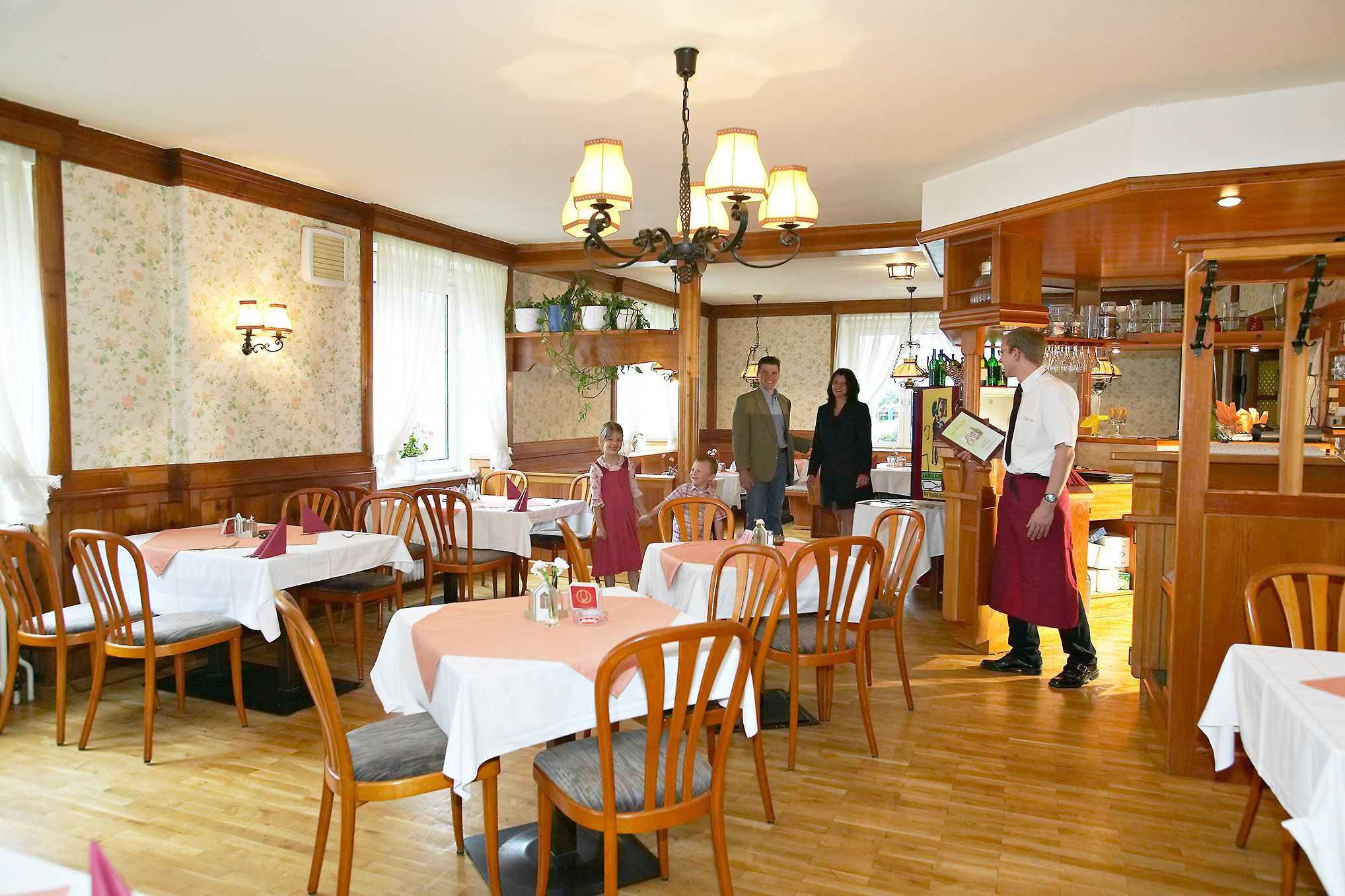 A rustic restaurant with wooden furniture and tablecloths. A waiter greets a family at the entrance.