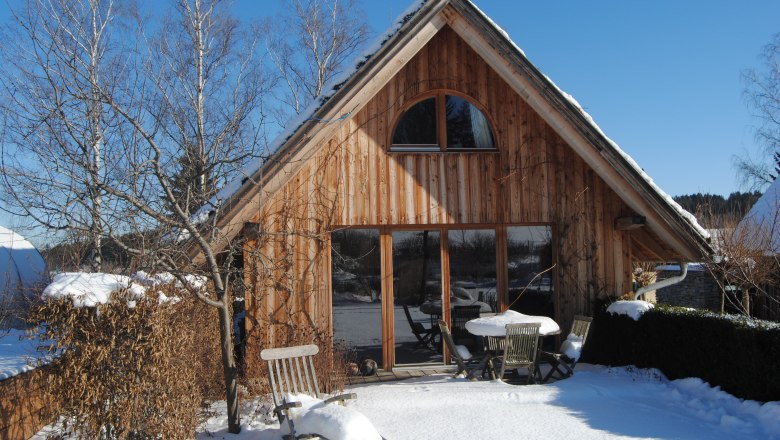 A wooden holiday home in the snow with garden furniture and bare trees.