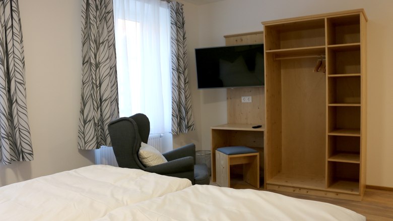 Open closet with TV on the wall. In front of a window with a curtain is a cozy green chair.