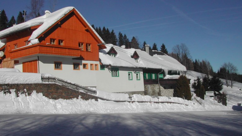 A snow-covered inn with a red wooden gable and white walls on the Feistritzsattel, surrounded by trees and a blue sky.