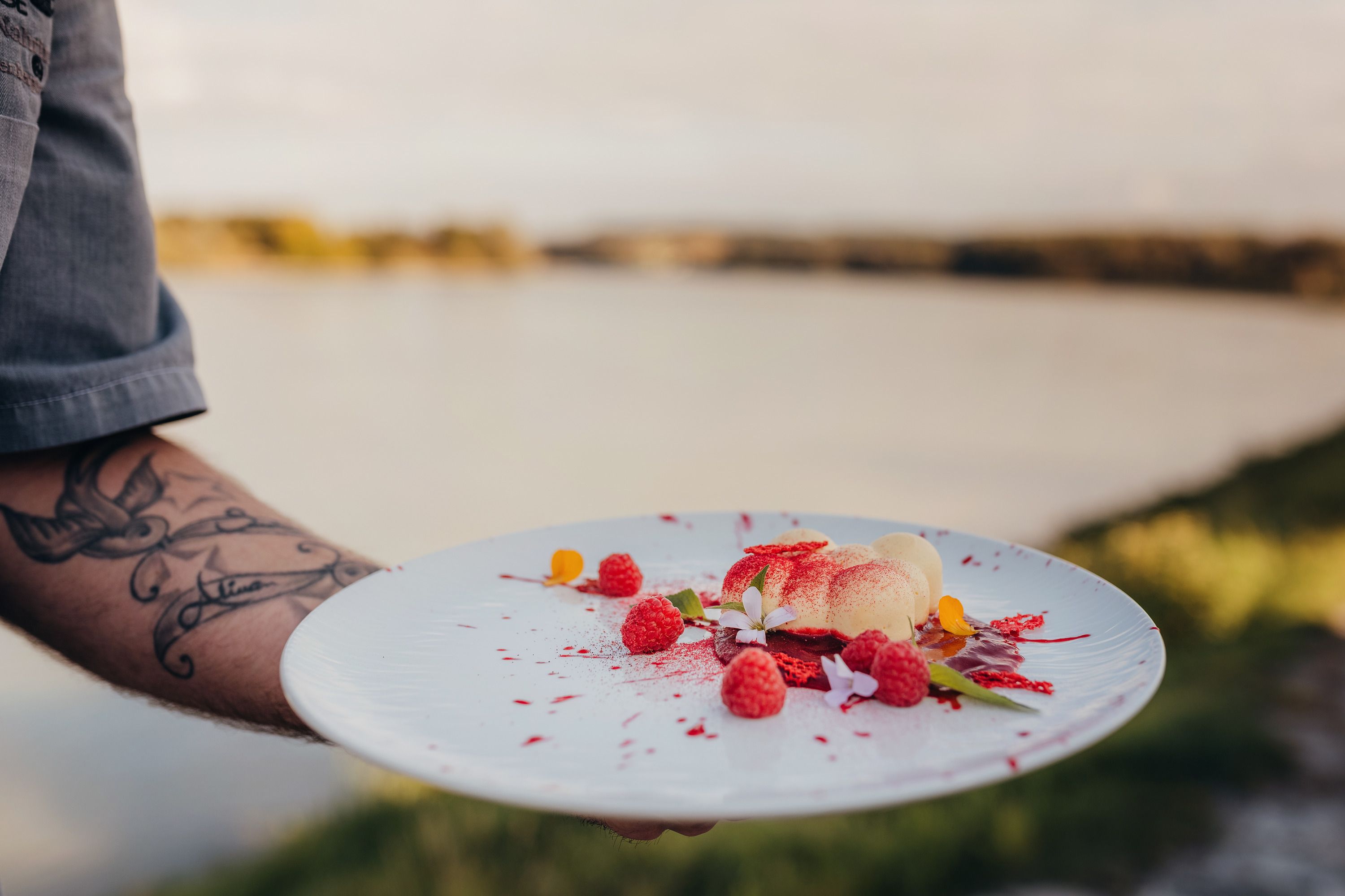 A plate of white chocolate mousse, raspberries and flowers, held by a tattooed hand in front of a lake.