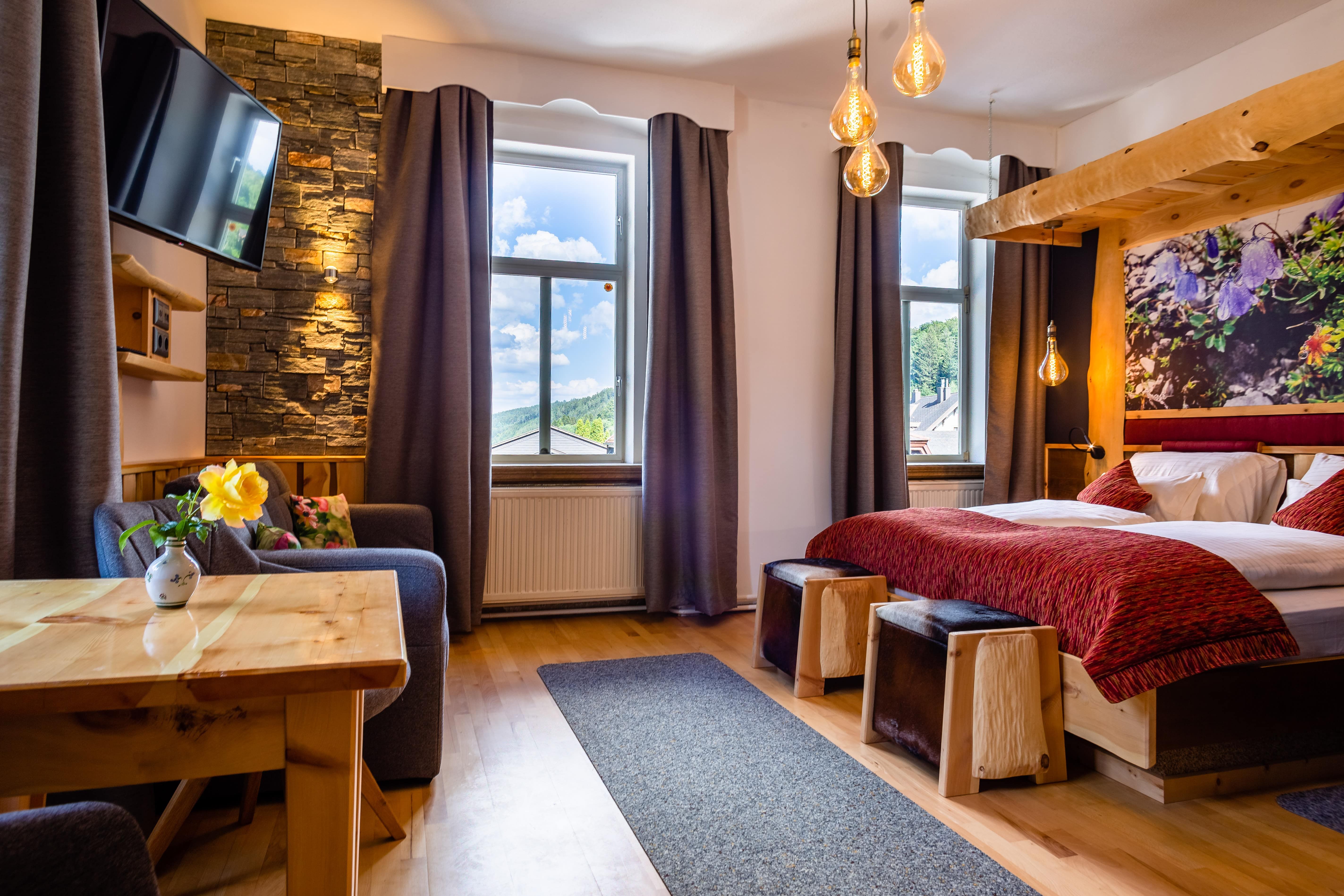 Cozy double room with wooden furniture, a bed, sofa, table and large windows with curtains.
