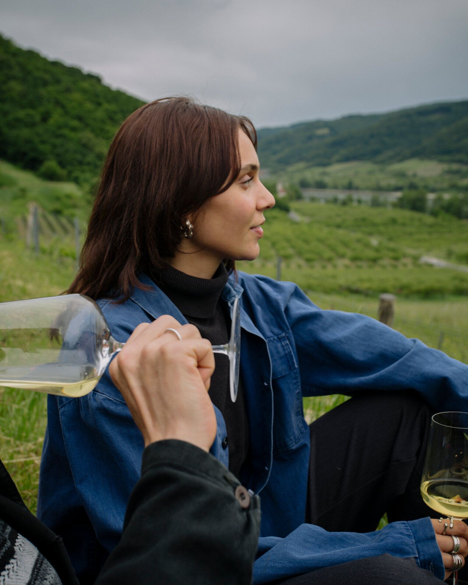Frau sitzt im Freien vor Weinbergen und Hügeln und blickt in die Landschaft; im Vordergrund halten zwei Personen Gläser mit Weißwein.