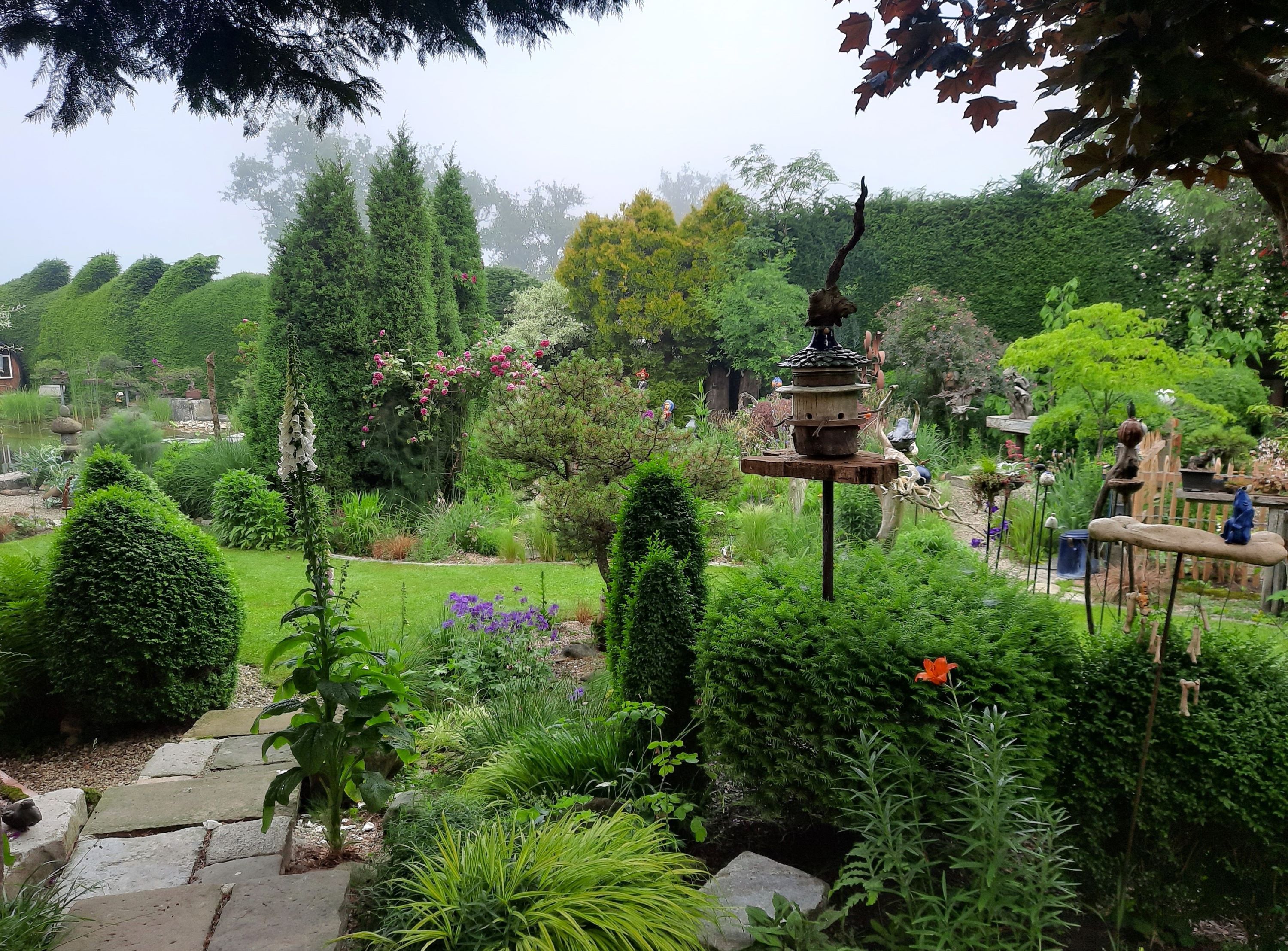 A lush garden with various plants, trees and decorations. A bird house stands in the foreground.