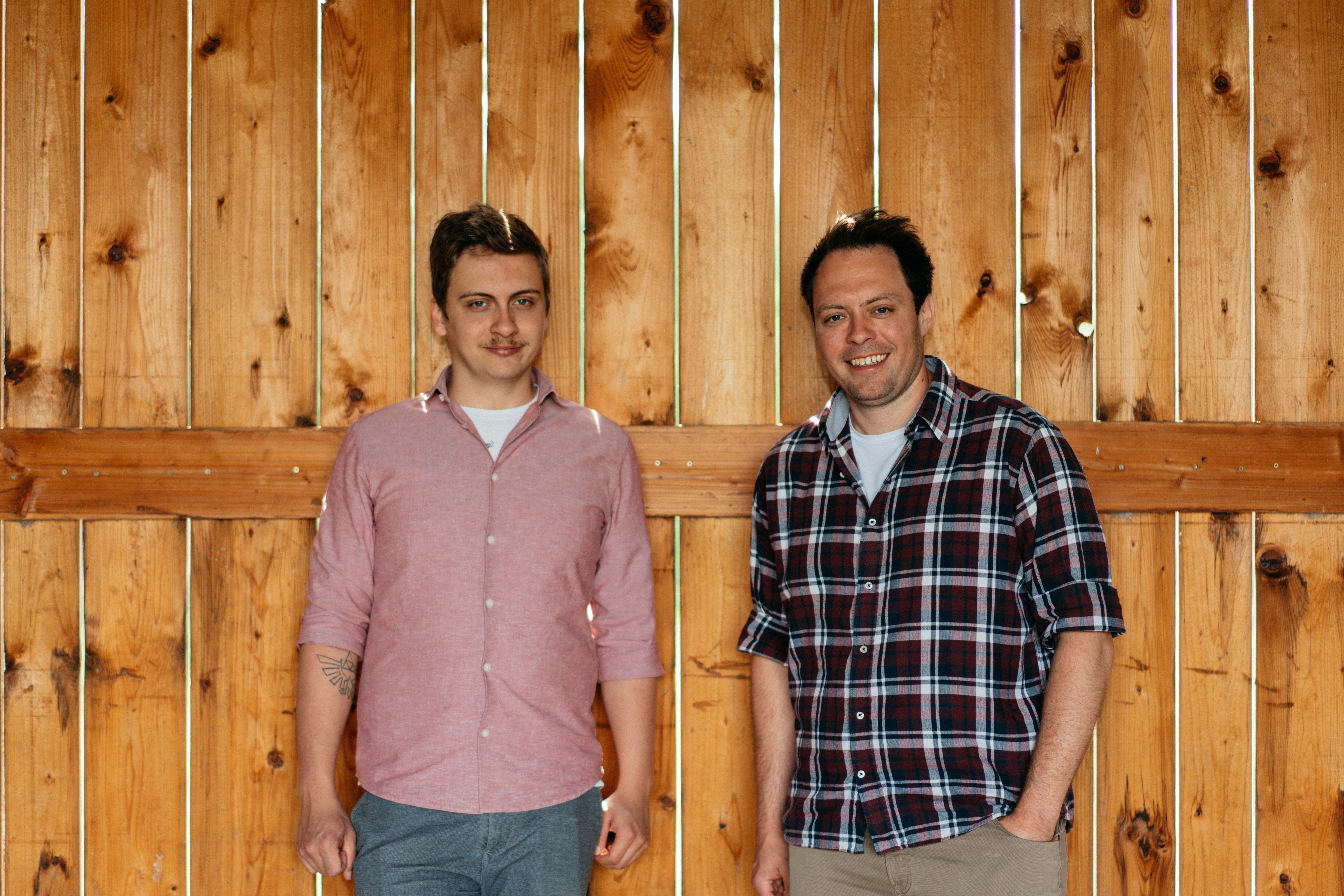 Two men stand in front of a wooden wall and smile into the camera.