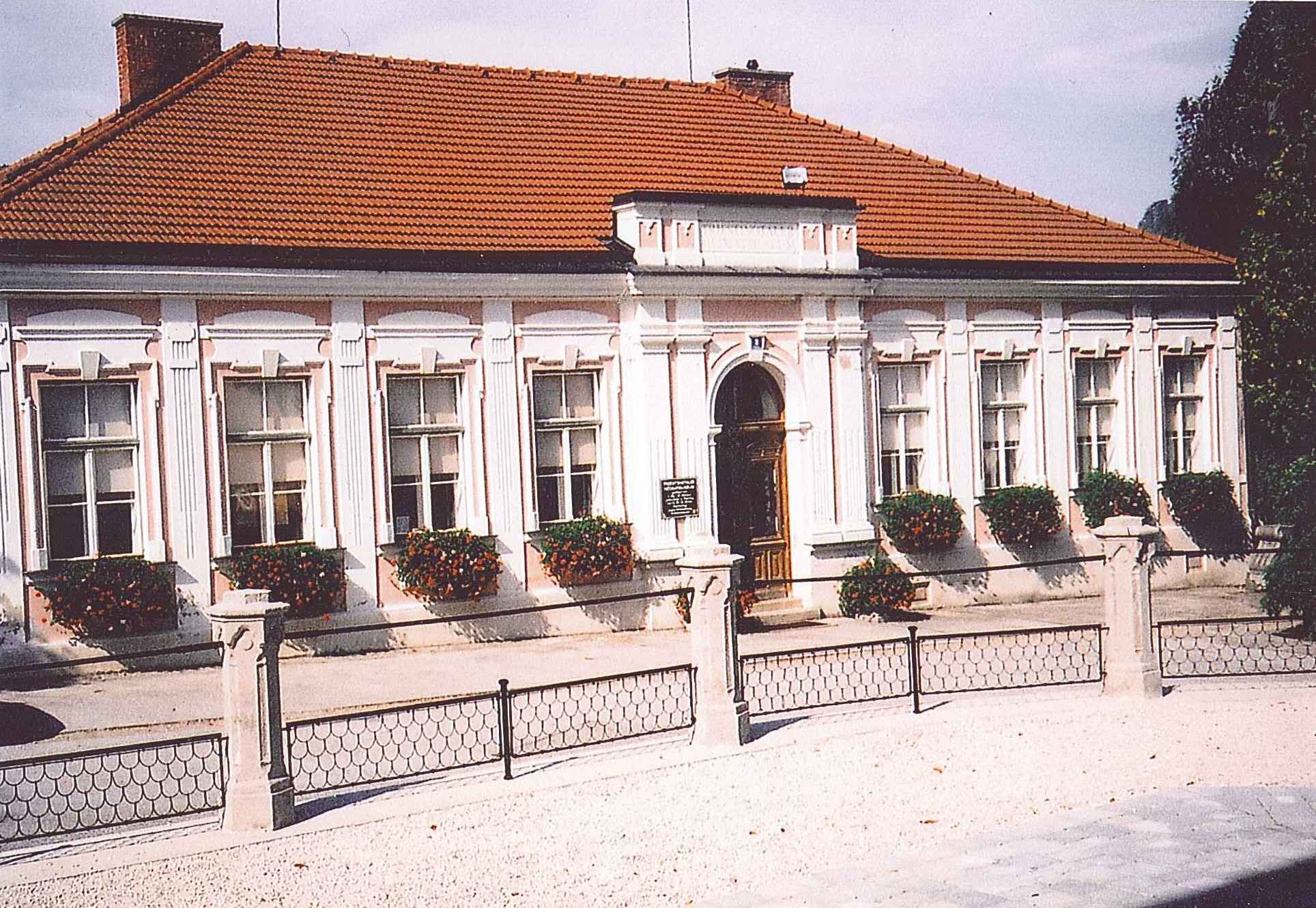 Historic building with red tiled roof and white window frames, surrounded by flowers.