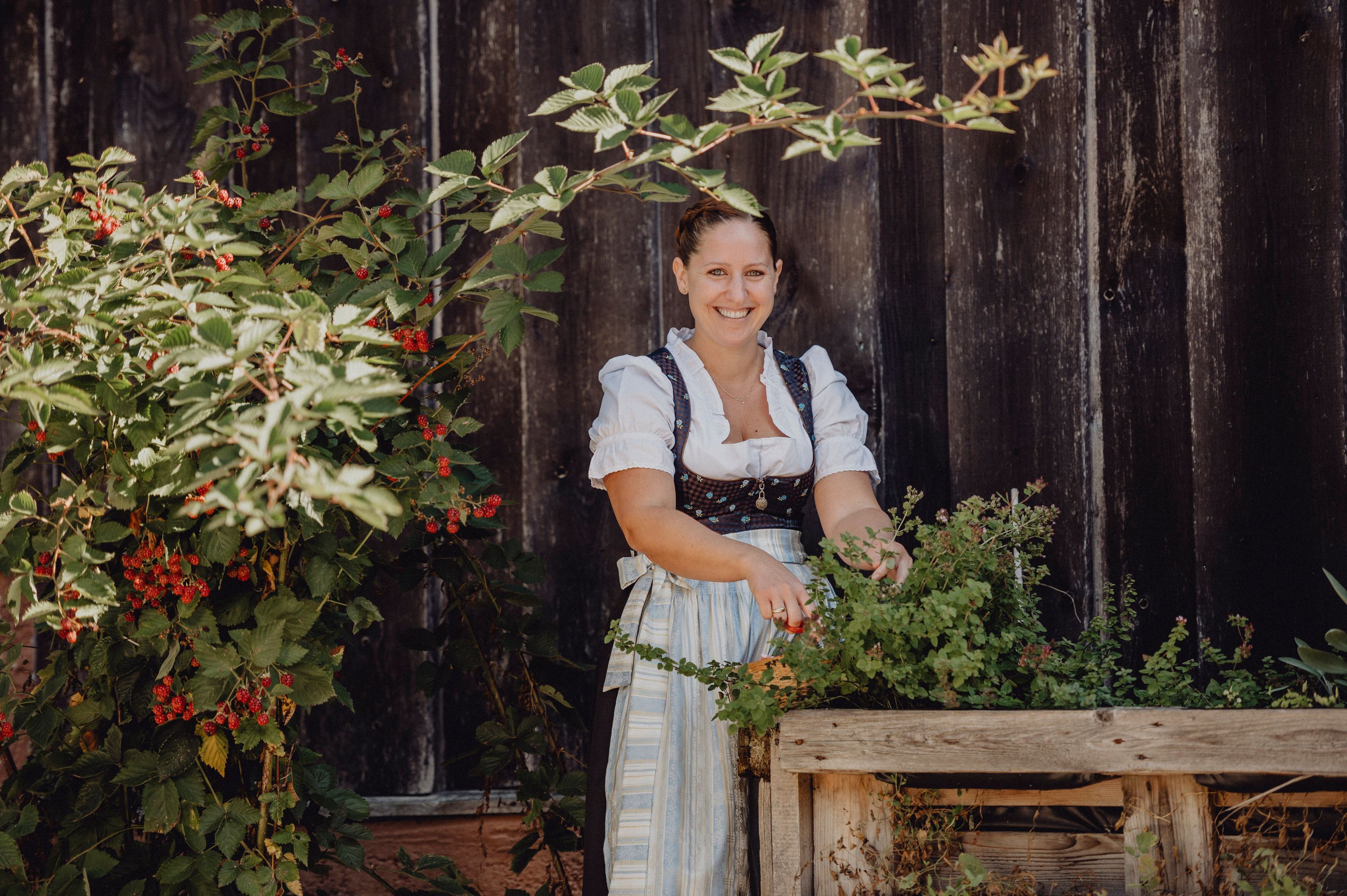 Woman in traditional dress stands smiling next to a bed of herbs in front of a wooden wall.