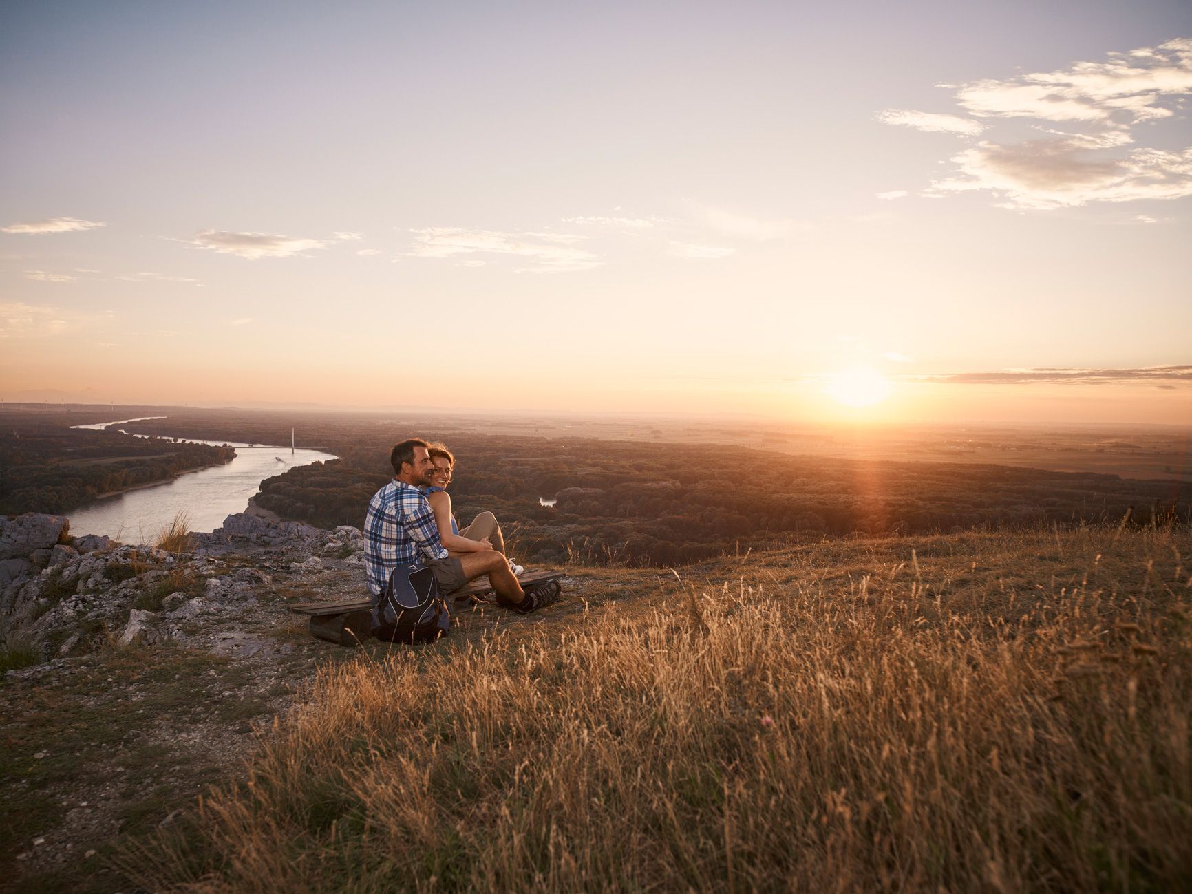 A couple sits on a hill and looks out over a river and the landscape at sunset.