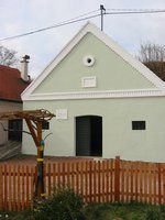A small, light green building with a pitched roof and a wooden fence in front of it.