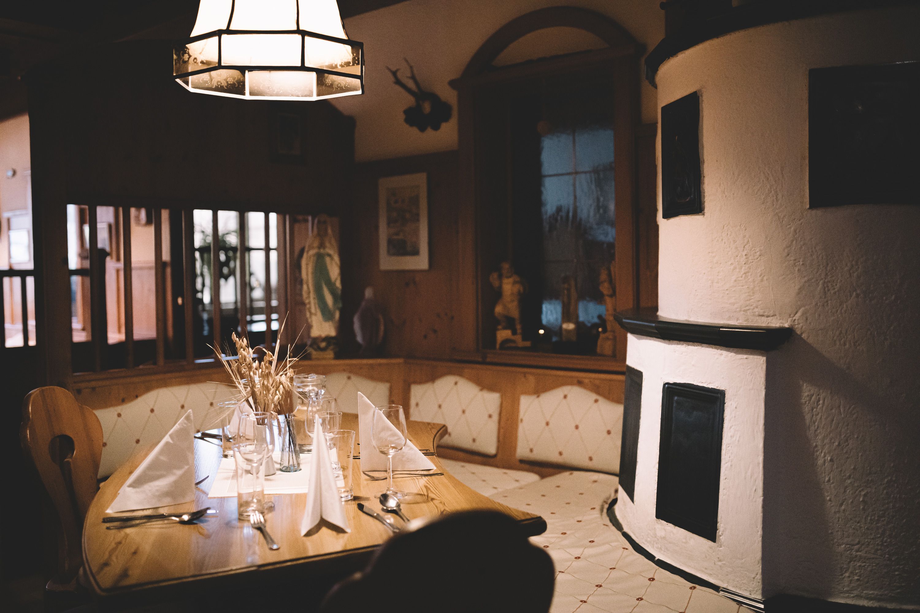 Cozy parlor with wooden table, set with glasses and napkins, next to a traditional tiled stove.