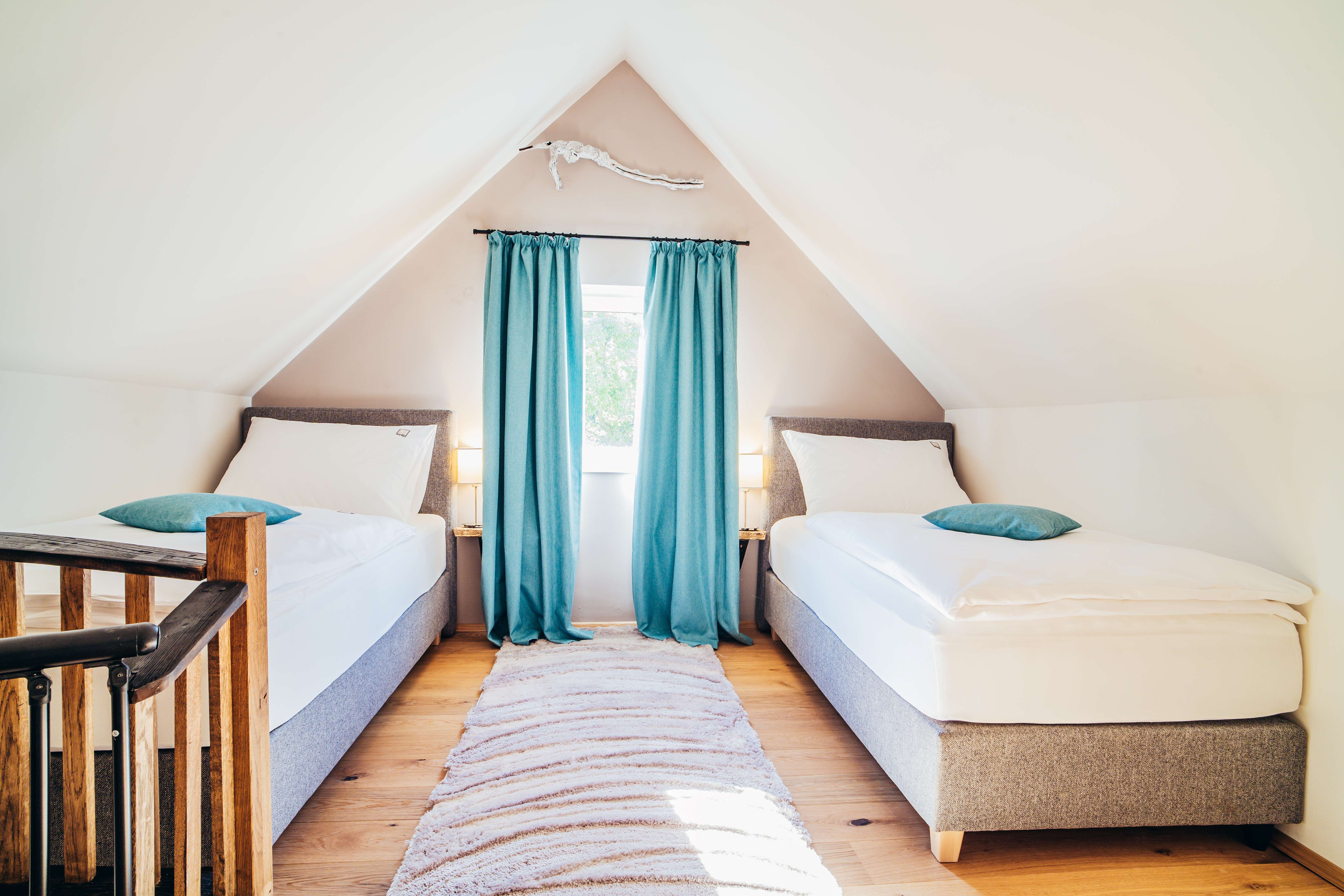 Two single beds in an attic room with blue curtains and wooden floor.