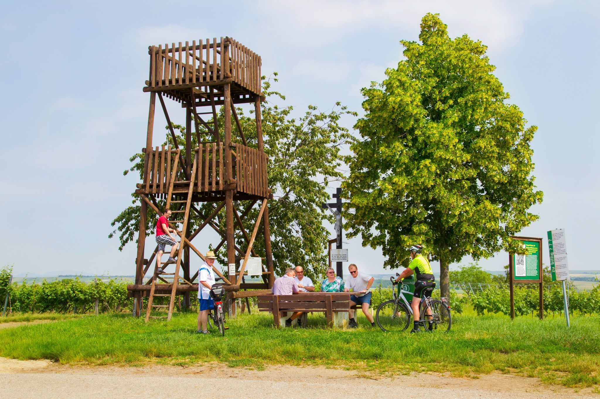 People relax at a wooden tower and table outside, surrounded by trees and signs.
