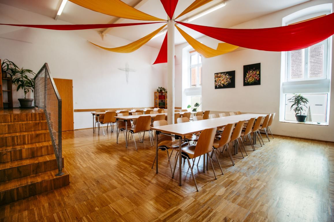 A bright dining room with a wooden floor, tables and chairs. Red and yellow fabric panels hang from the ceiling.