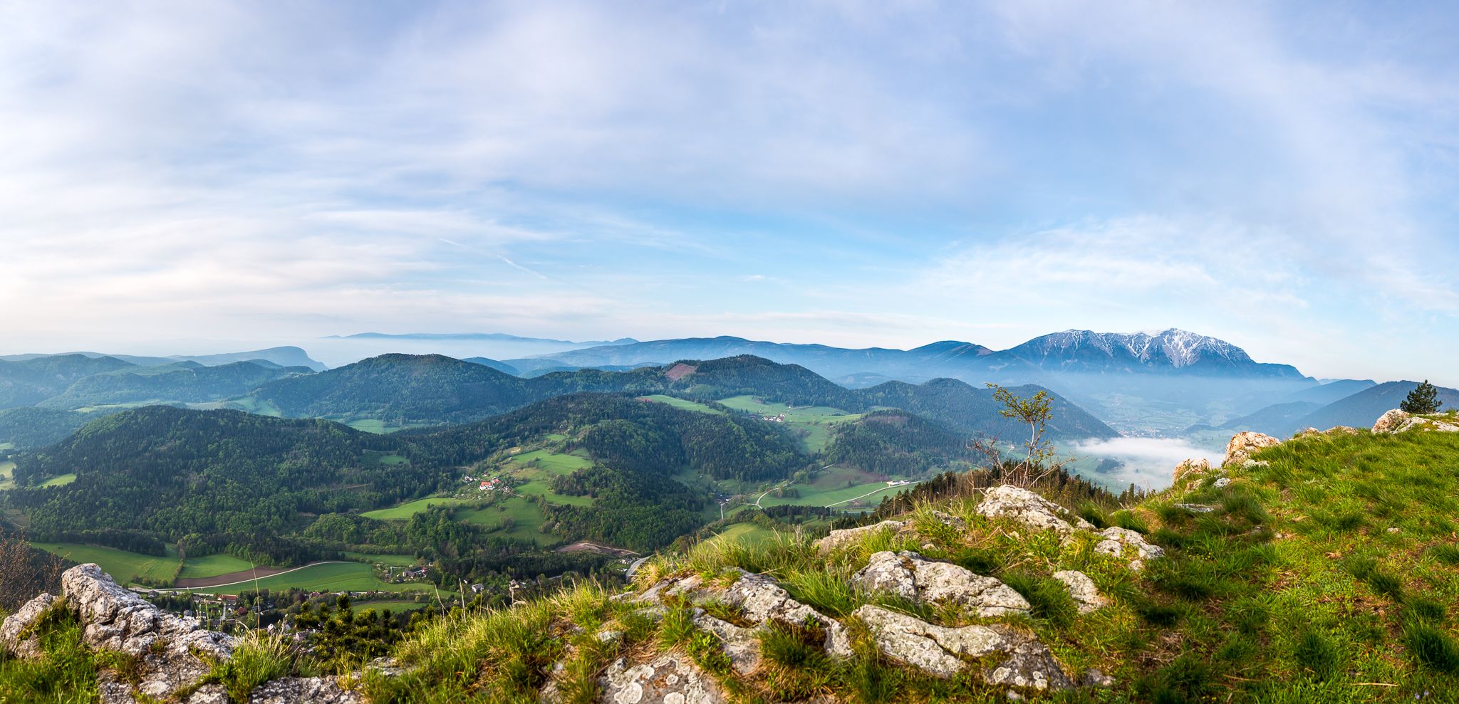 Panoramic view of a green mountain landscape with wooded hills and a snow-capped mountain in the background under a blue sky.