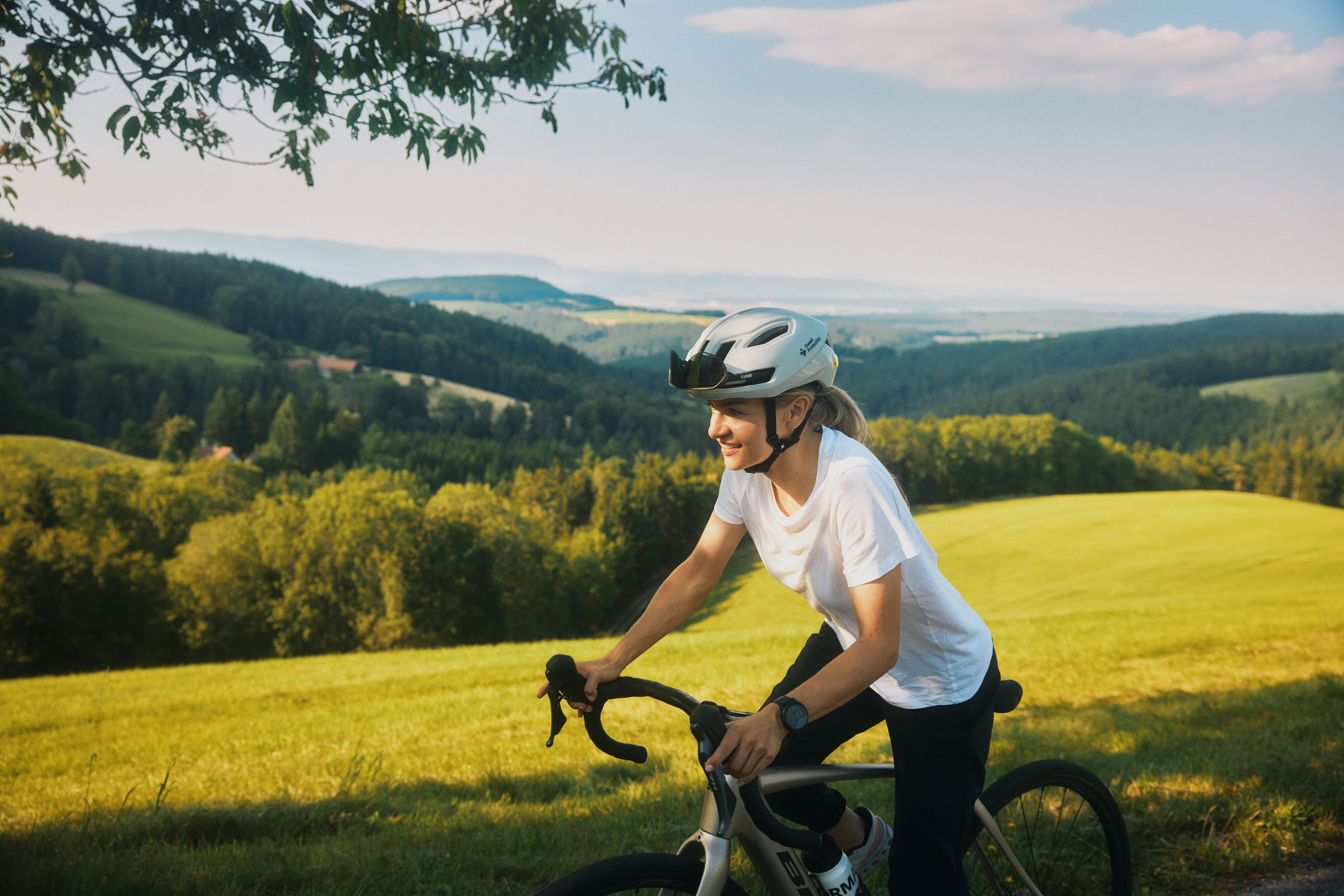 A person rides a bicycle through a hilly landscape with green meadows and forests in the background.