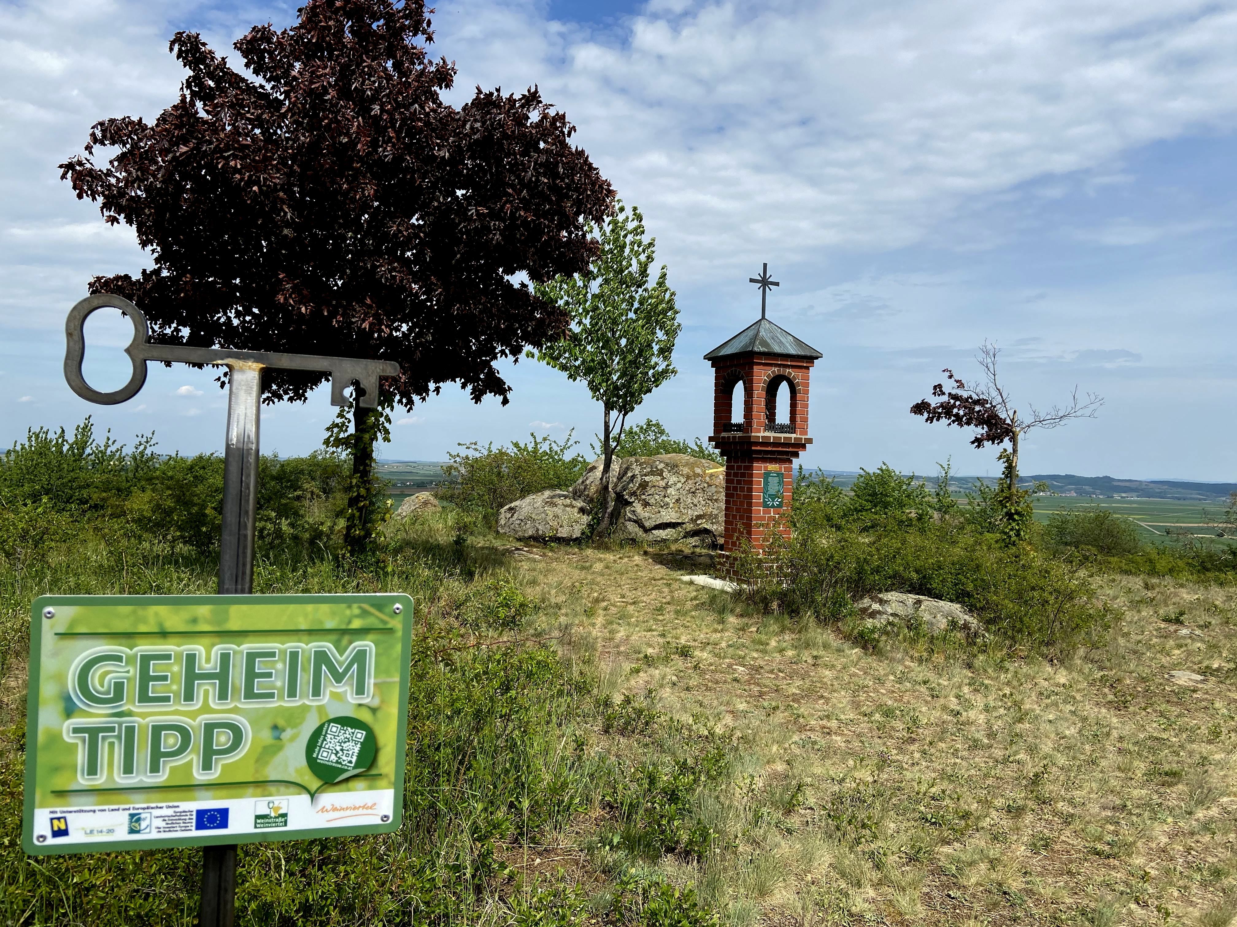 A sign with the inscription 'Geheim Tipp' in front of a small brick tower with a cross on a hill.