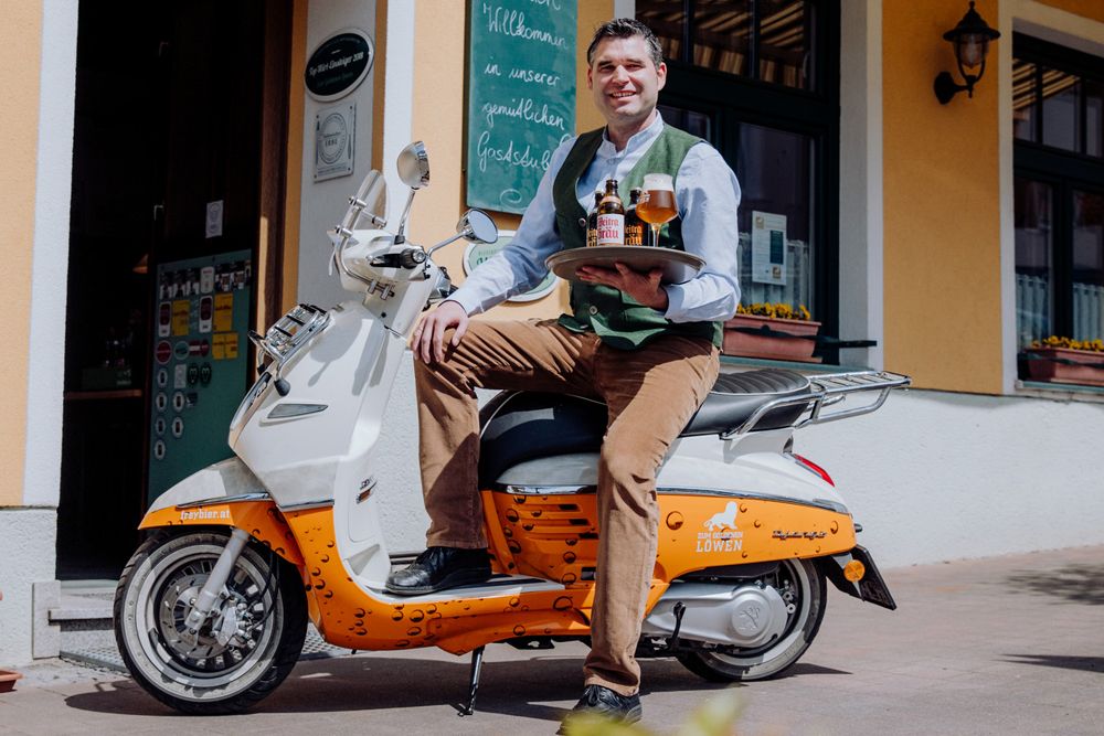A man sits on a scooter in front of a building holding a tray with beer bottles and a glass.
