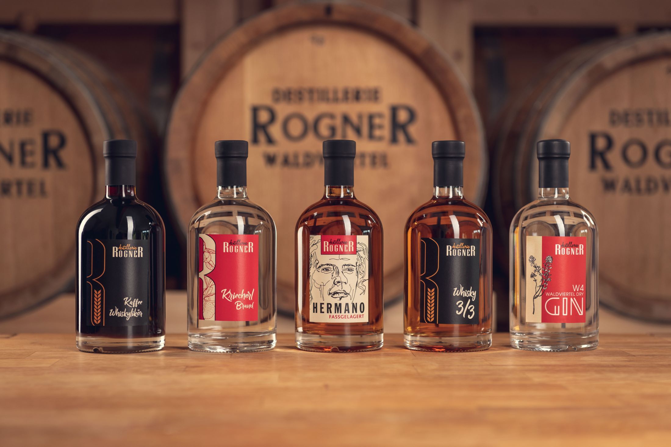 Five bottles of spirits from the Rogner distillery in front of wooden barrels.