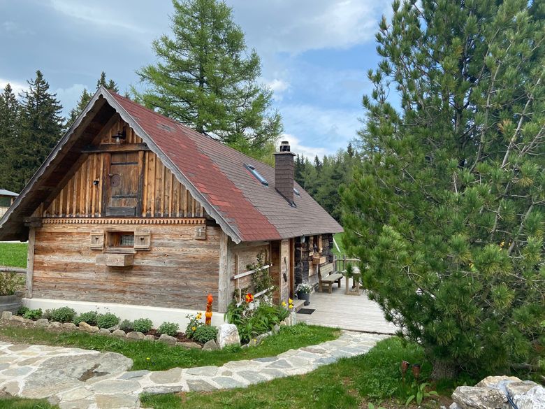 A rustic wooden hut with a red roof in a wooded setting, surrounded by trees and a well-tended garden.