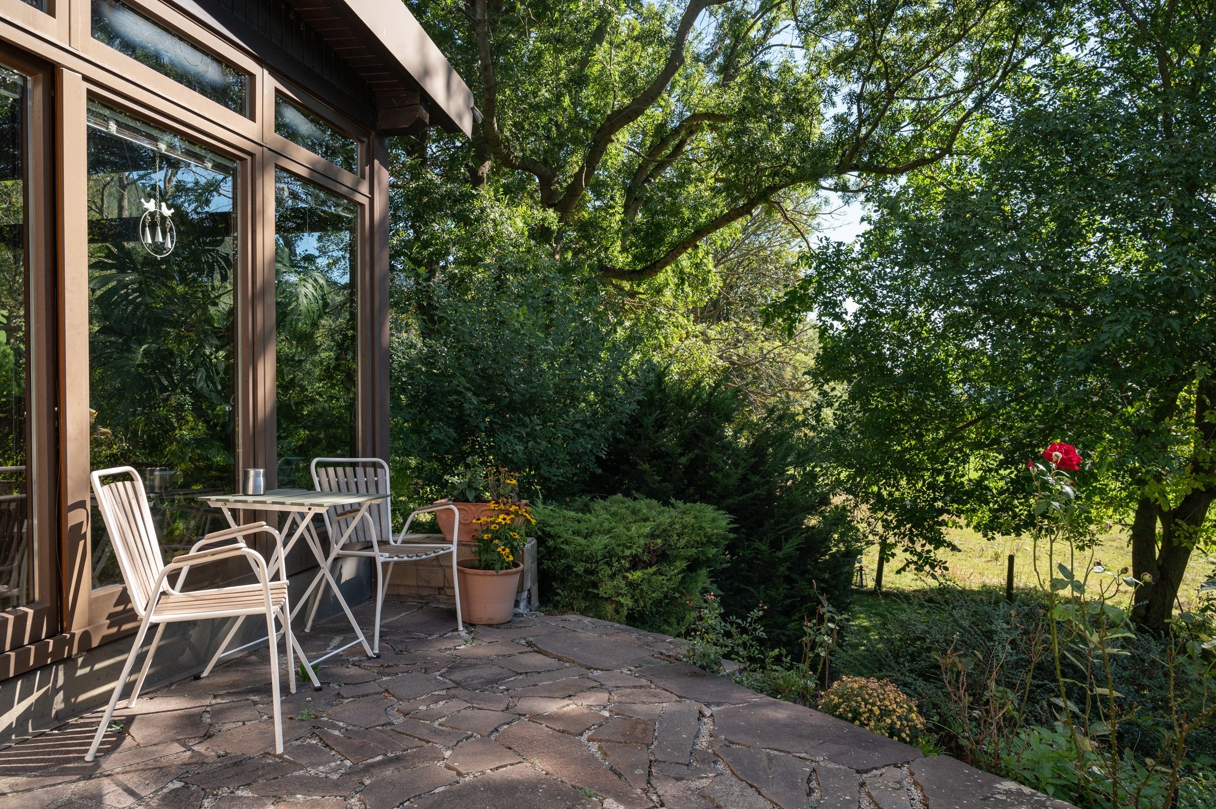 Terrace with table and chairs in front of a vacation home, surrounded by trees and plants.