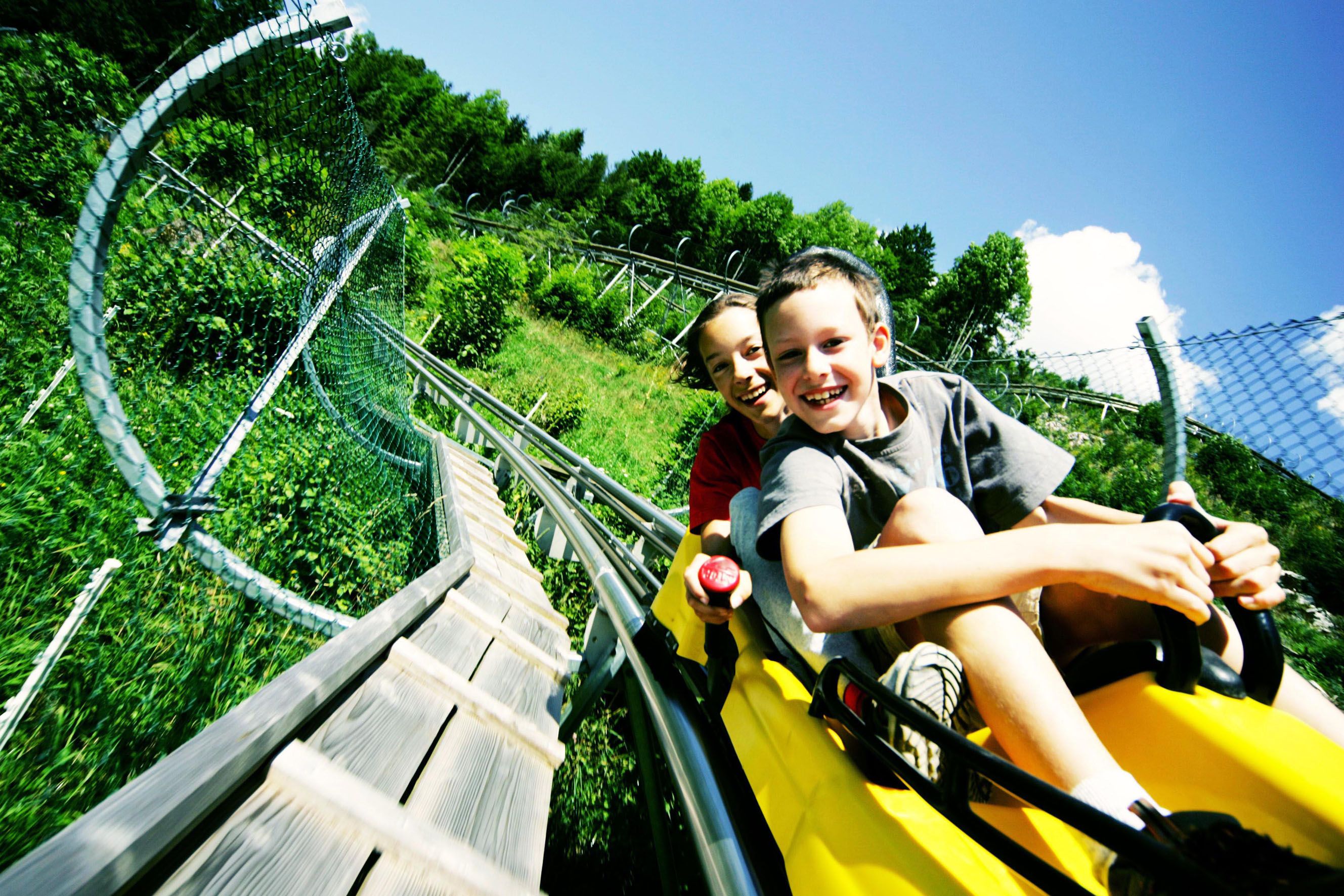 Two children laughing on a summer toboggan run through a green landscape.