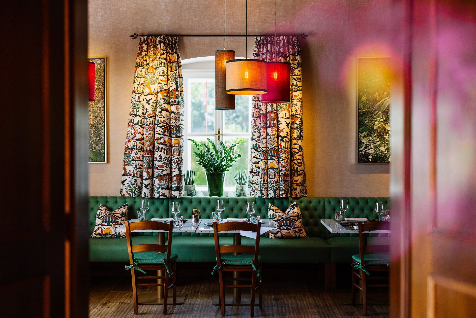 Cozy dining area with large windows, green upholstered furniture and plants, with a view of the countryside.