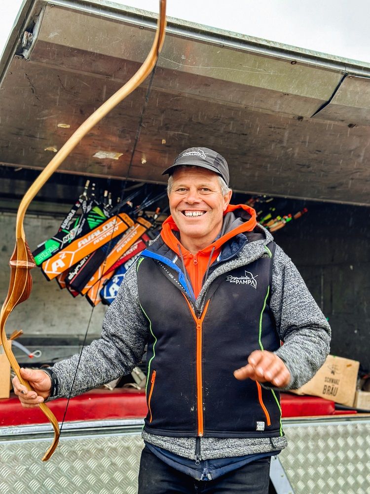 A smiling man holds a bow in front of a pendant with arrows.
