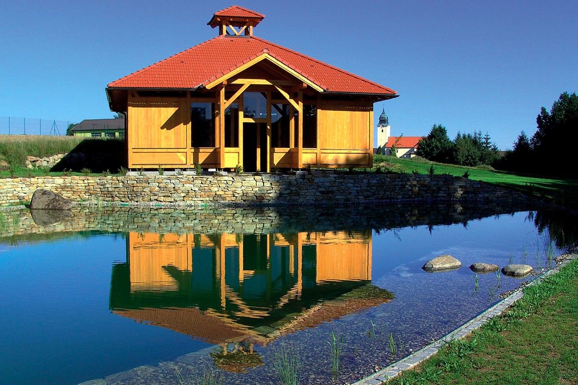 A wooden building with a red roof is reflected in a pond surrounded by a green landscape.