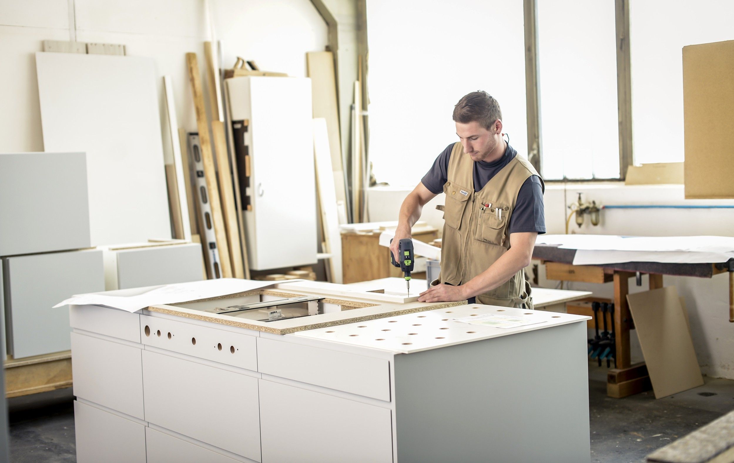A craftsman is working on a piece of furniture in a workshop with a cordless screwdriver.